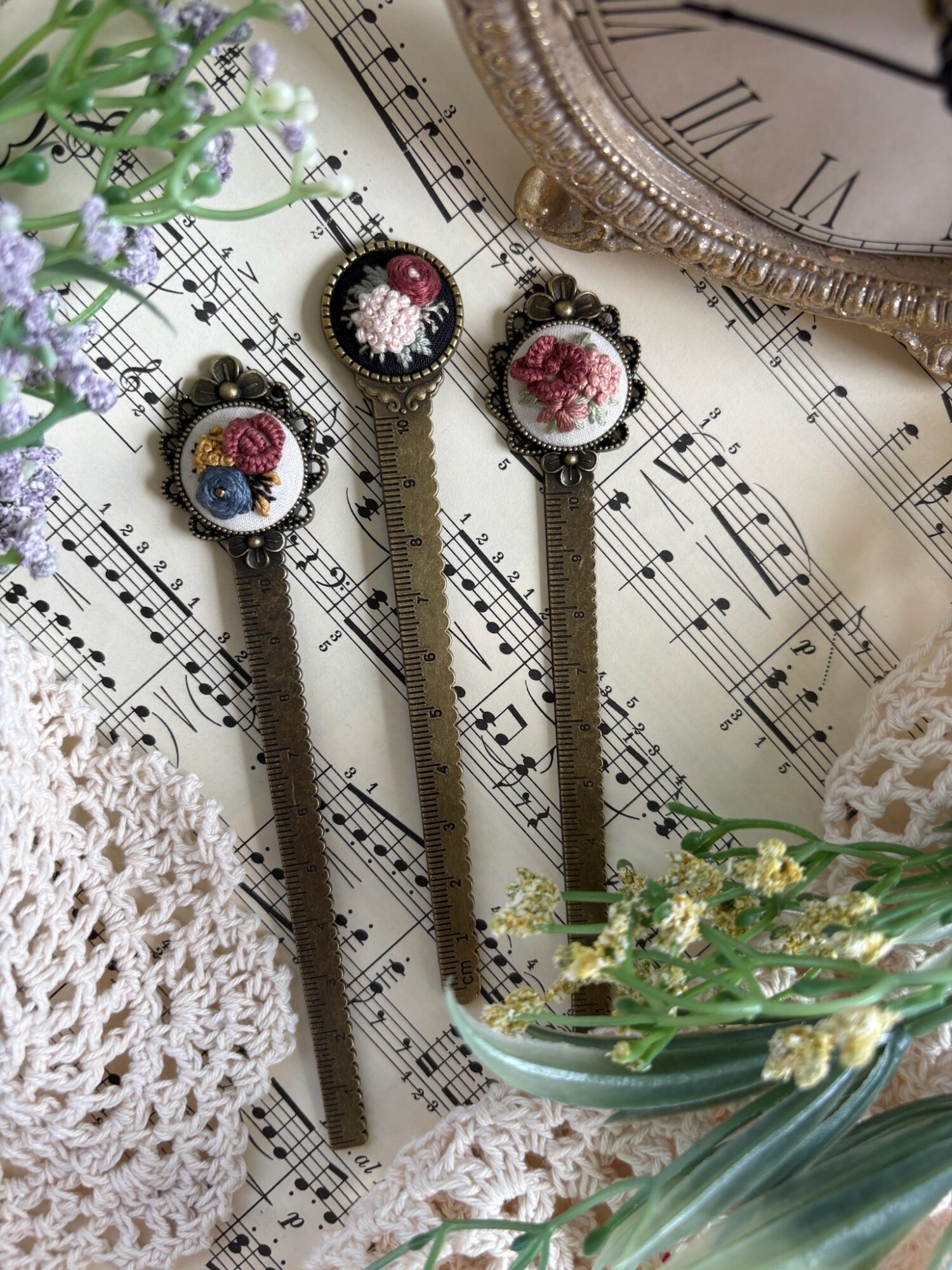 Three vintage-style pocket watches with floral designs on the covers, placed on sheet music with a clock and crochet item nearby.