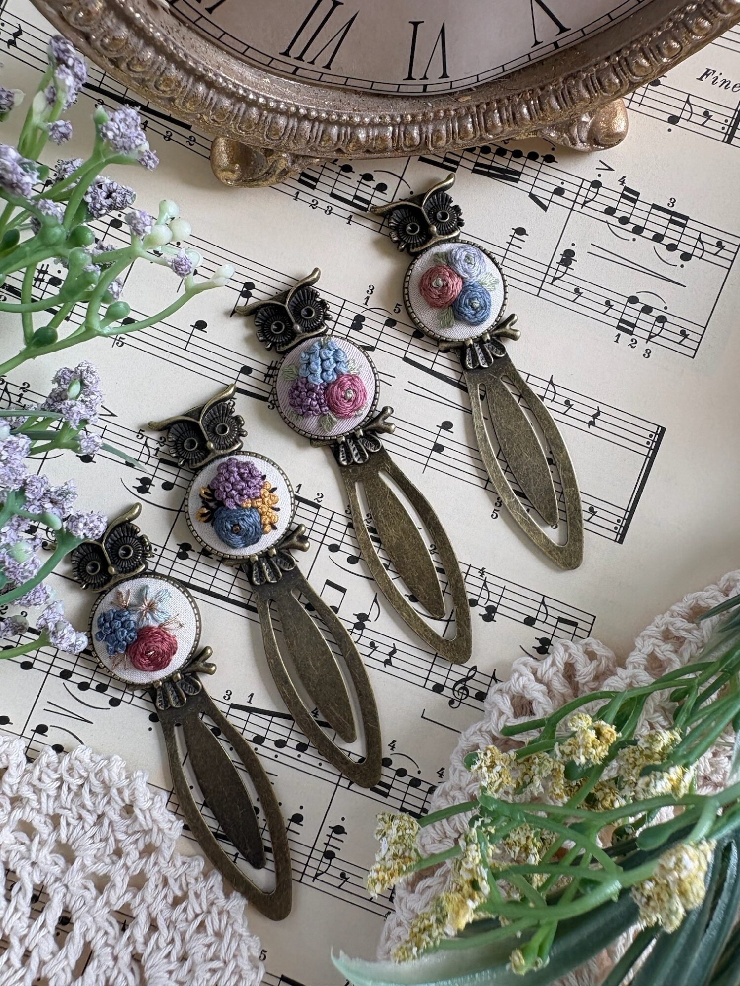 Two pairs of vintage-style earrings with floral and oval designs, placed on sheet music with flowers and a clock nearby.