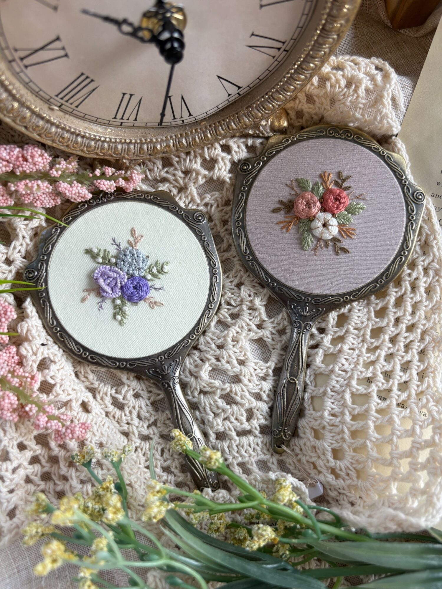 Two vintage hand mirrors with floral embroidery on lace fabric, surrounded by pink and yellow flowers, and a clock in the background.