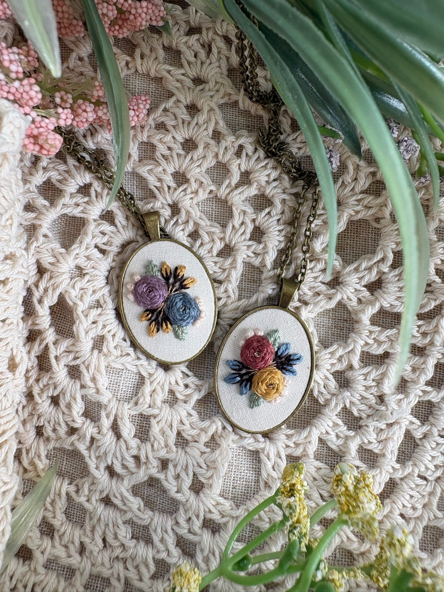 Two oval earrings with floral designs, one with purple and yellow flowers, the other with pink and yellow flowers, on a crochet surface.