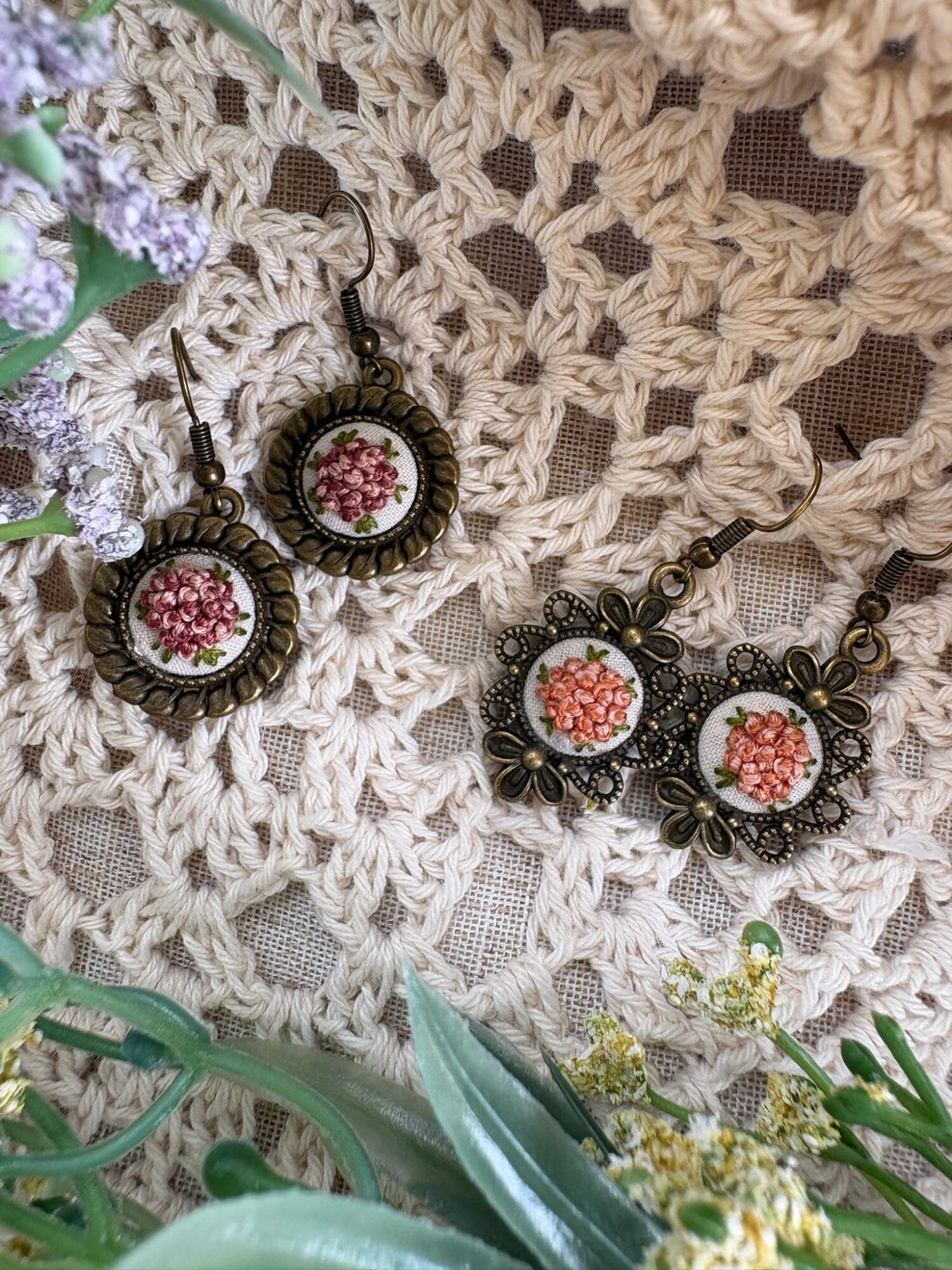 Pair of vintage-style earrings with floral embroidery, placed on a crocheted fabric with flowers around.
