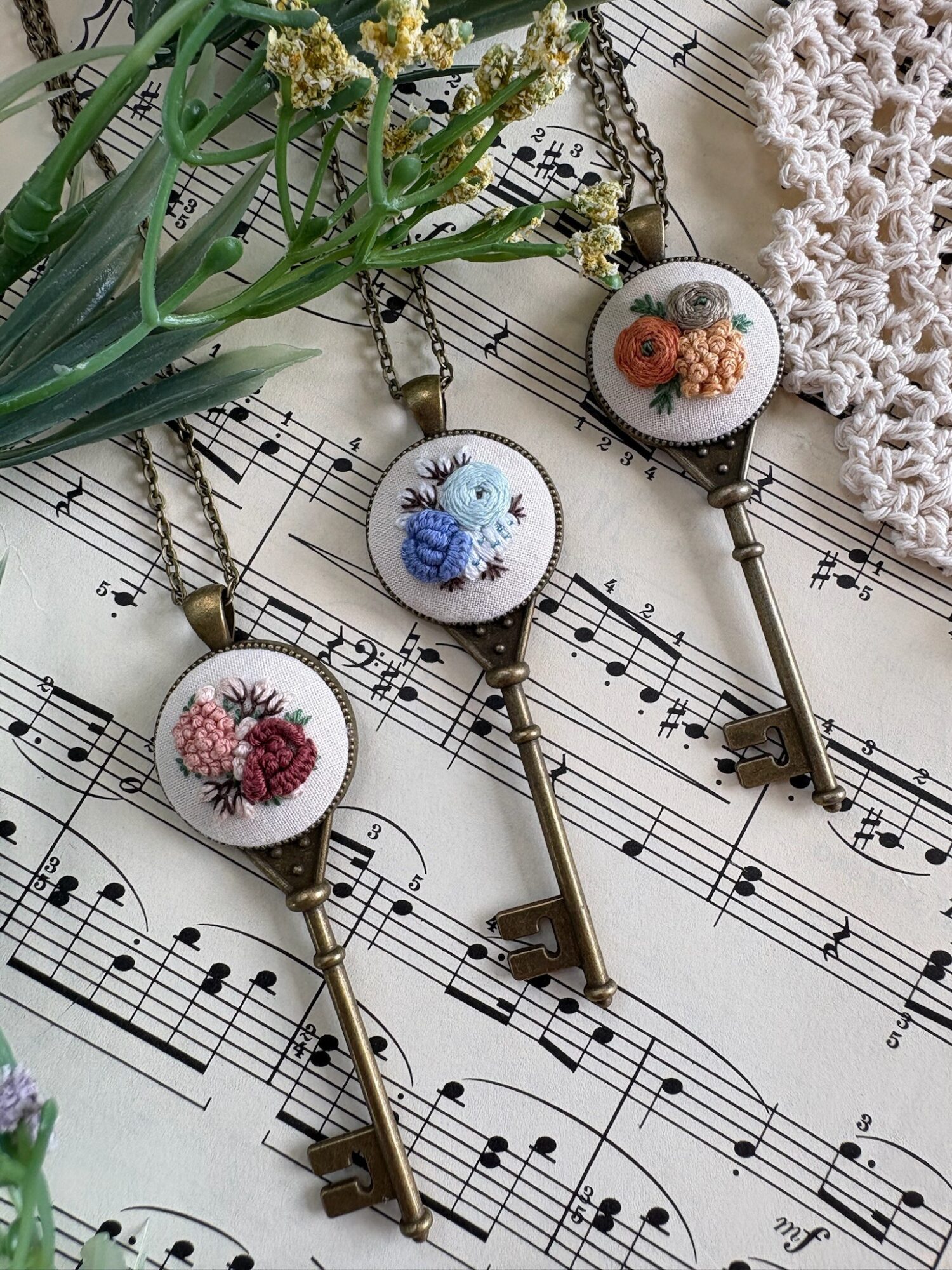 Three vintage-style keys with round floral embroidery tops, placed on sheet music with a lace cloth and flowers nearby.