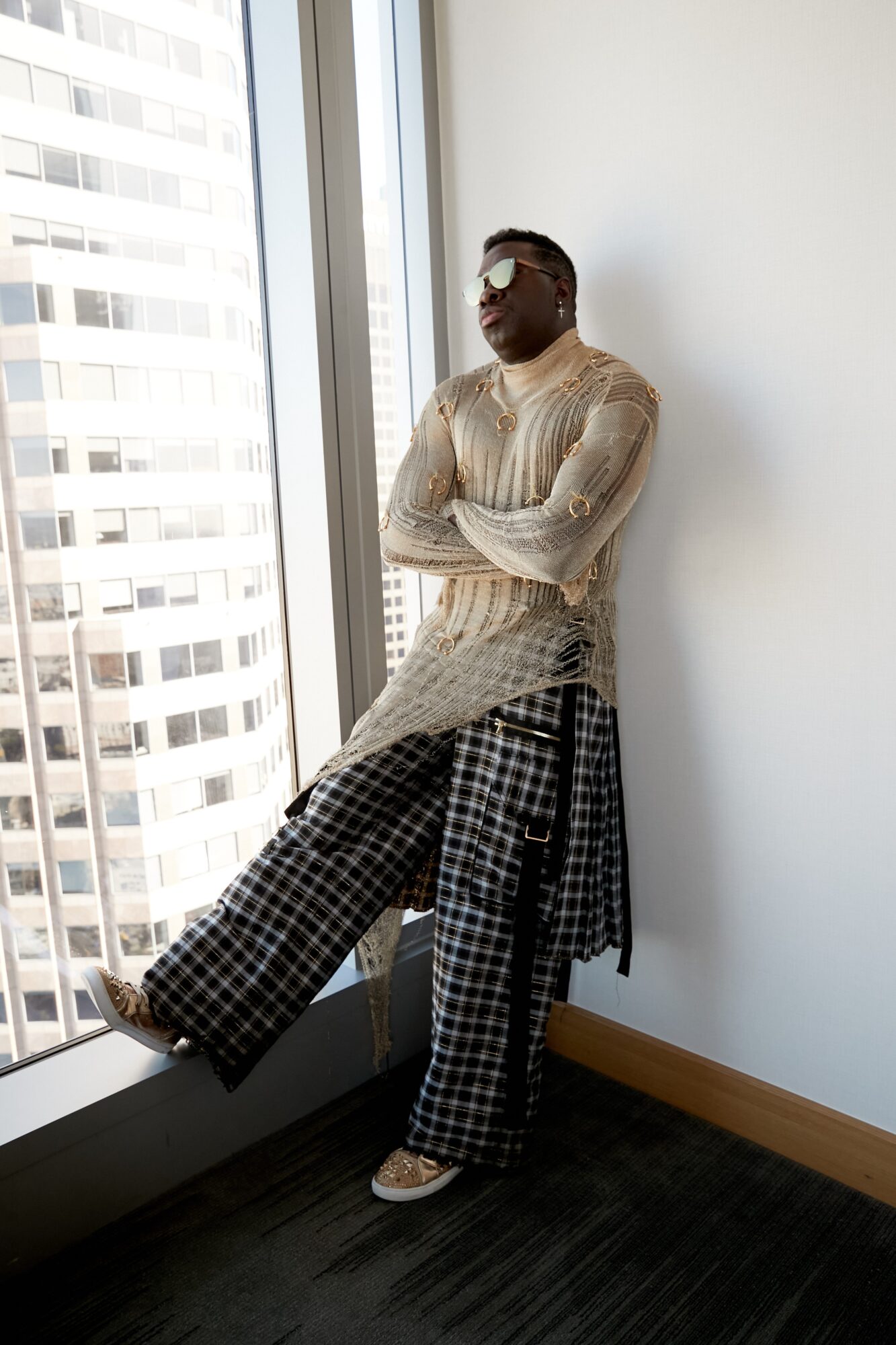 Person standing by window with arms crossed, wearing sunglasses, a beige sweater, and checkered pants, in a bright room.