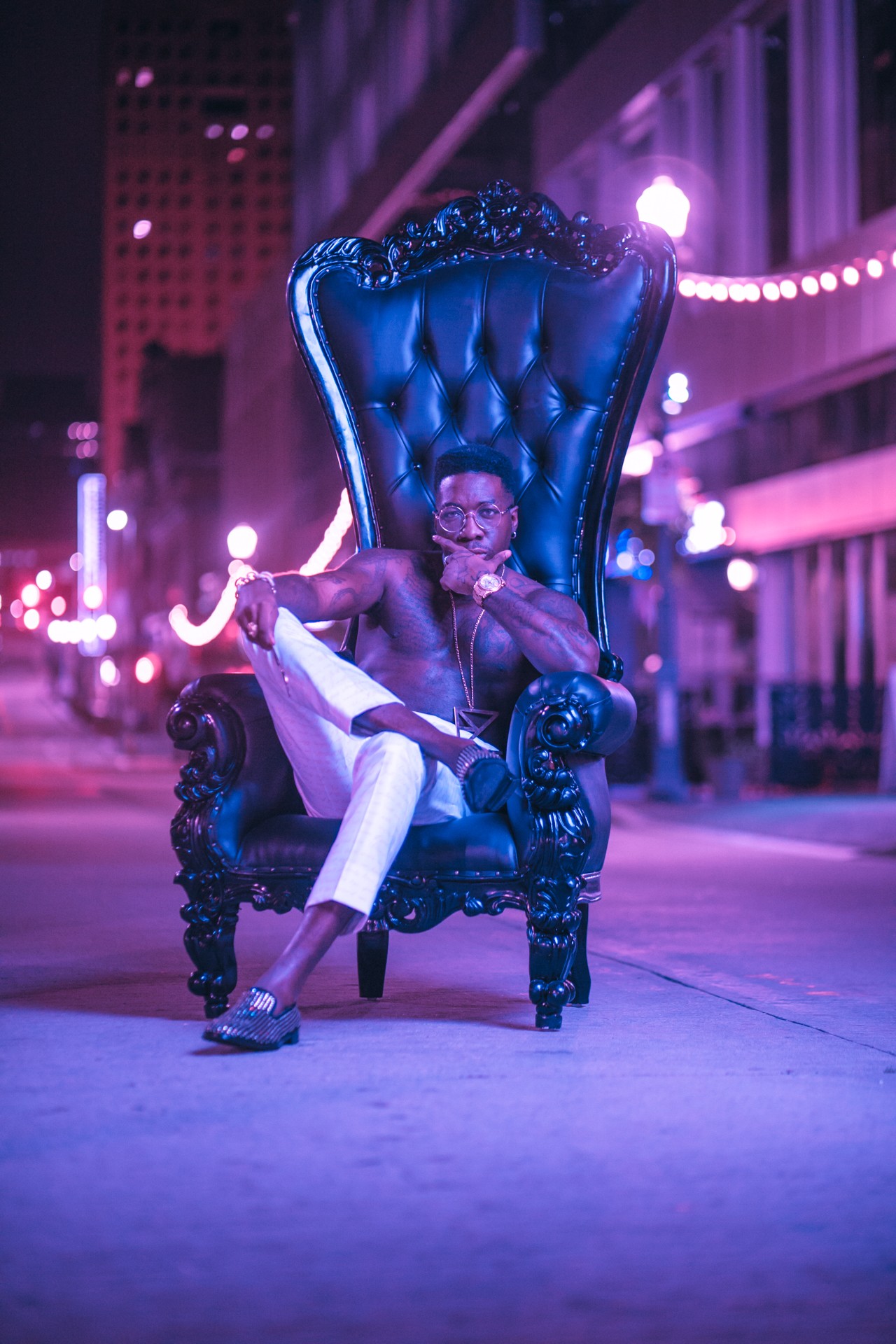 Person sitting on a large ornate blue throne outdoors at night, illuminated by purple and pink city lights.