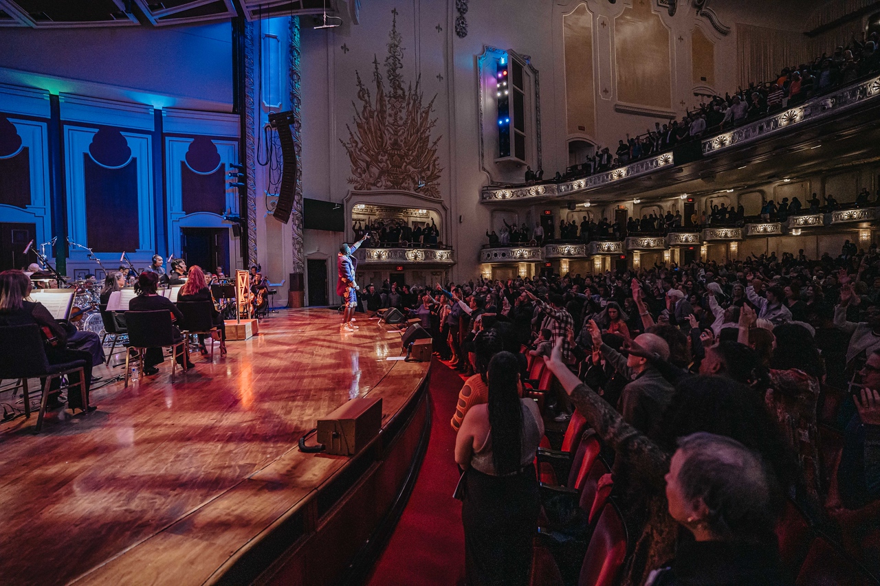 Concert hall with stage, audience seated, and ornate interior decor, illuminated with blue and warm lighting.