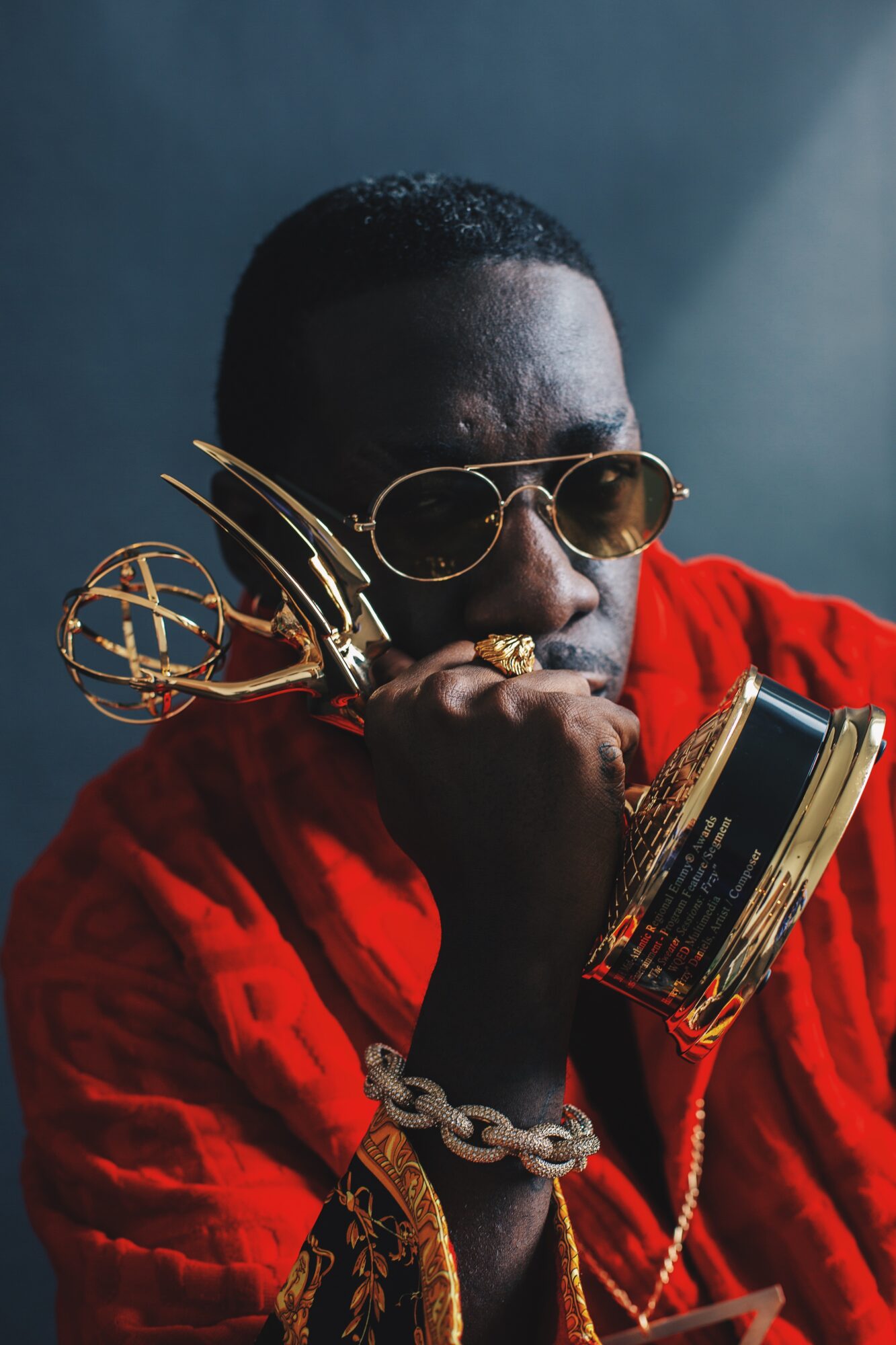 Person with sunglasses and jewelry, wearing a red garment, resting chin on hand, against dark background.