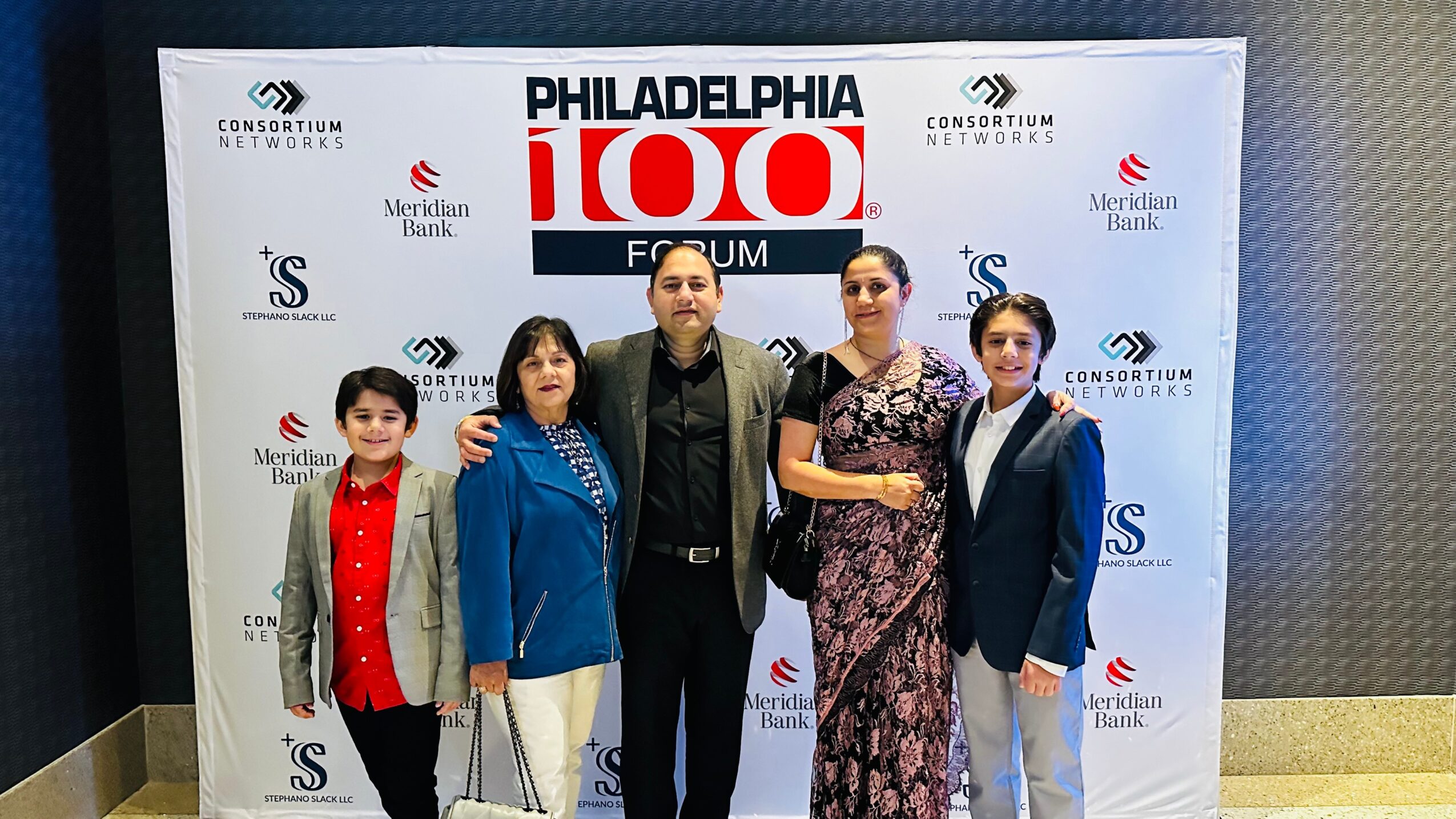 Group of five people standing in front of a Philadelphia 100 forum backdrop, smiling, with two women and three children, in formal and casual attire.