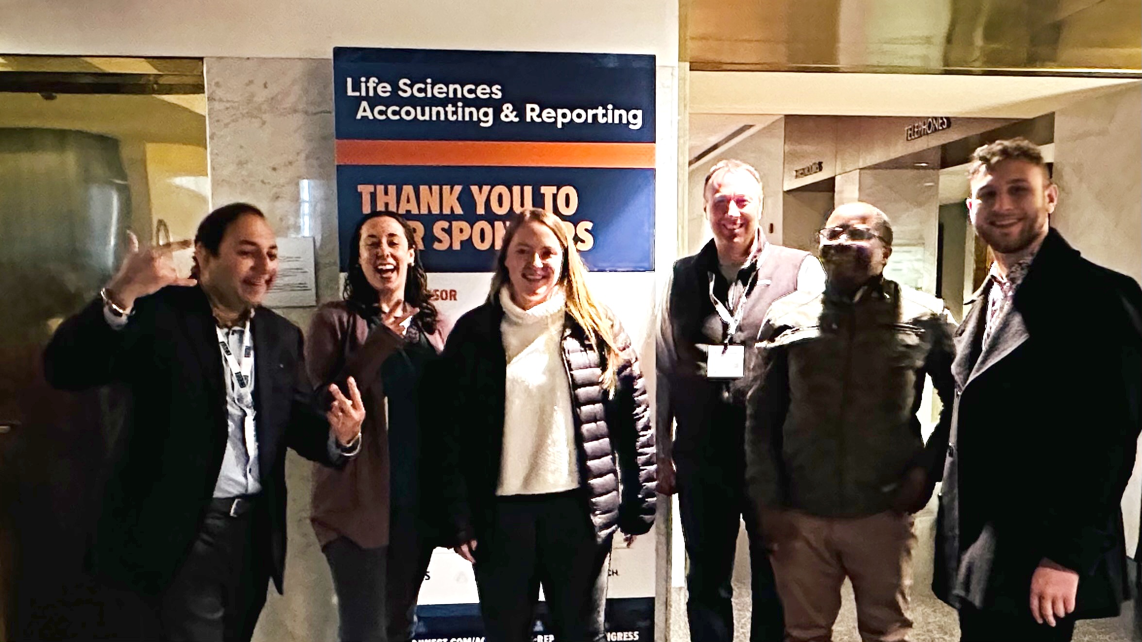 Group of six people standing together indoors, smiling, with a sign behind them that reads 'Life Sciences Accounting & Reporting'.