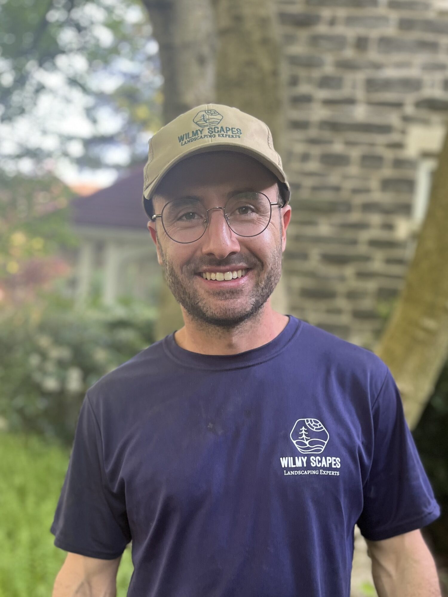 Man wearing glasses, beige cap, and navy T-shirt outdoors with trees and stone wall in background.
