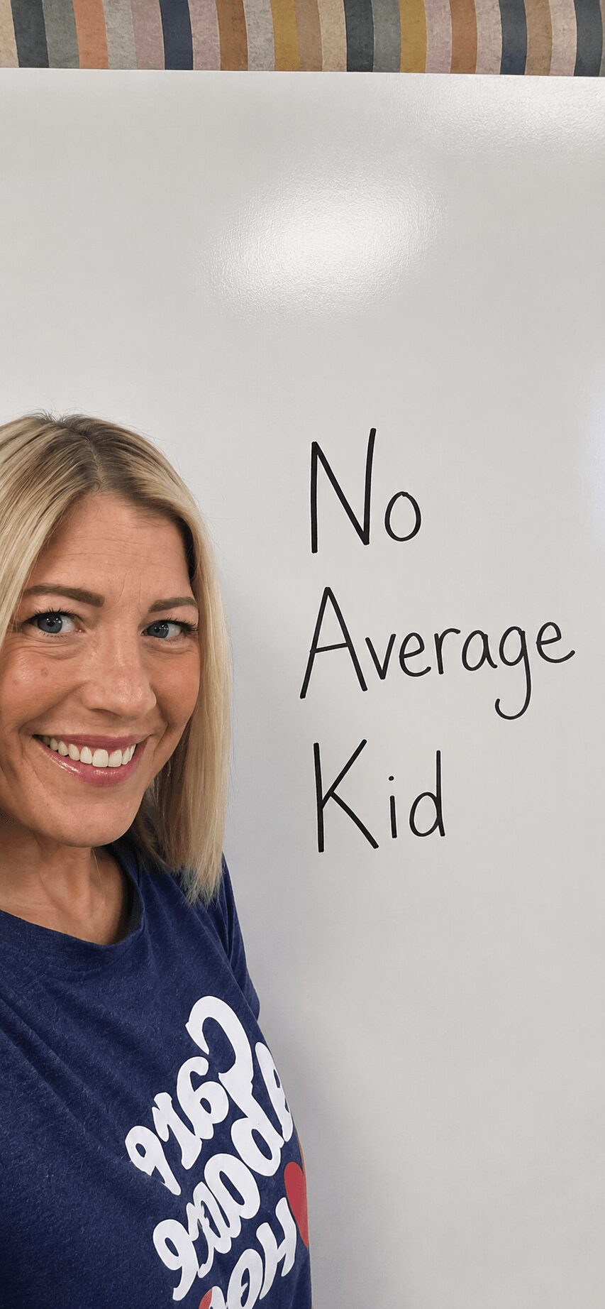 Smiling woman with blonde hair wearing a blue shirt, standing next to a whiteboard with the words 'No Average Kid'.