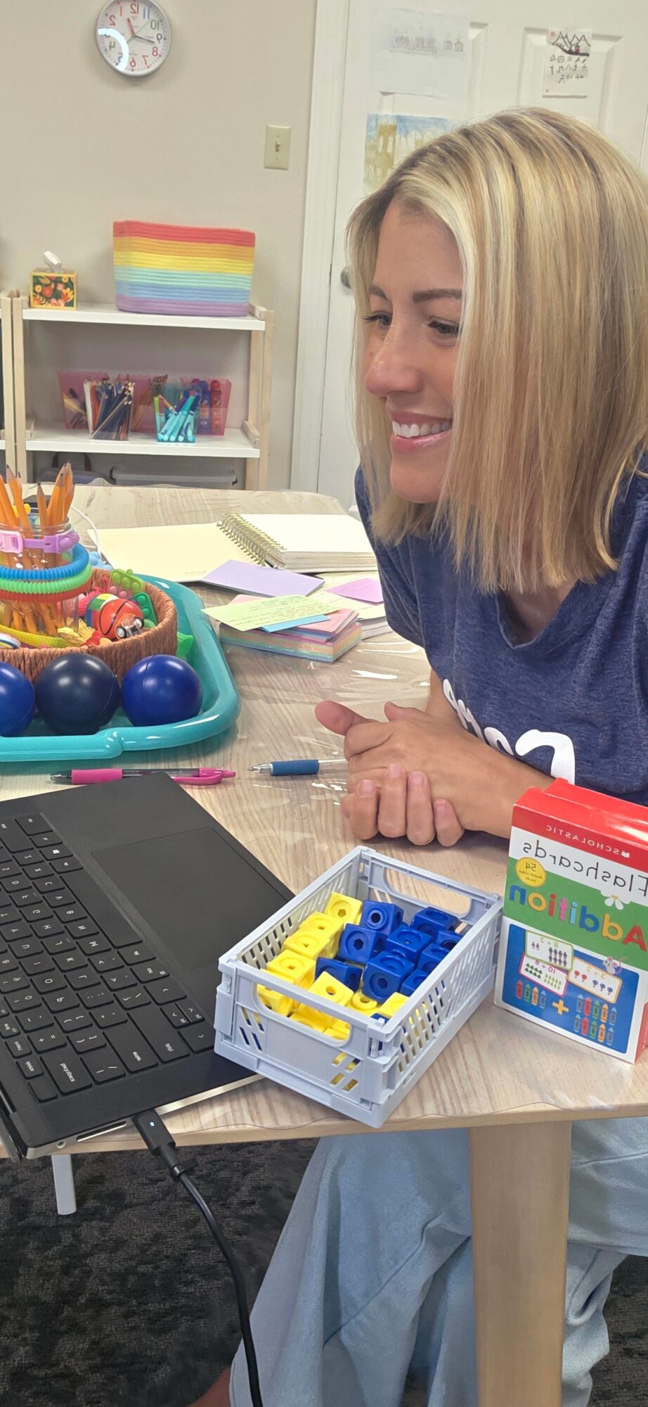 Woman with blonde hair smiling at a desk with colorful toys and a laptop, in a classroom setting.