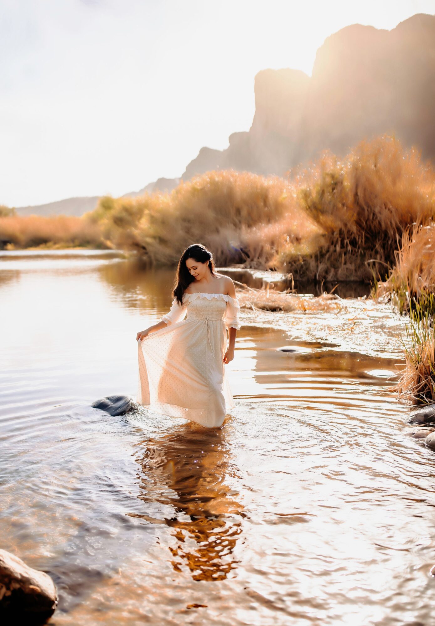 Woman in a white dress standing in shallow water near a riverbank with mountains and bushes in the background, sunlight shining brightly.