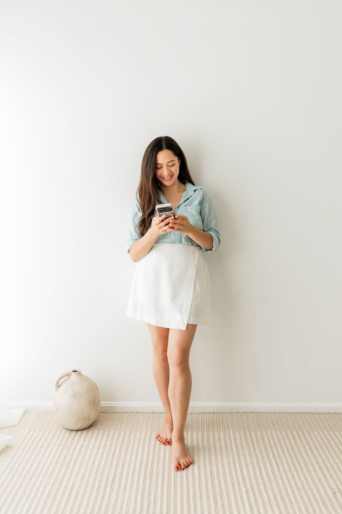 Young woman standing barefoot, looking at her phone, wearing a denim jacket and white dress, against a plain white wall.