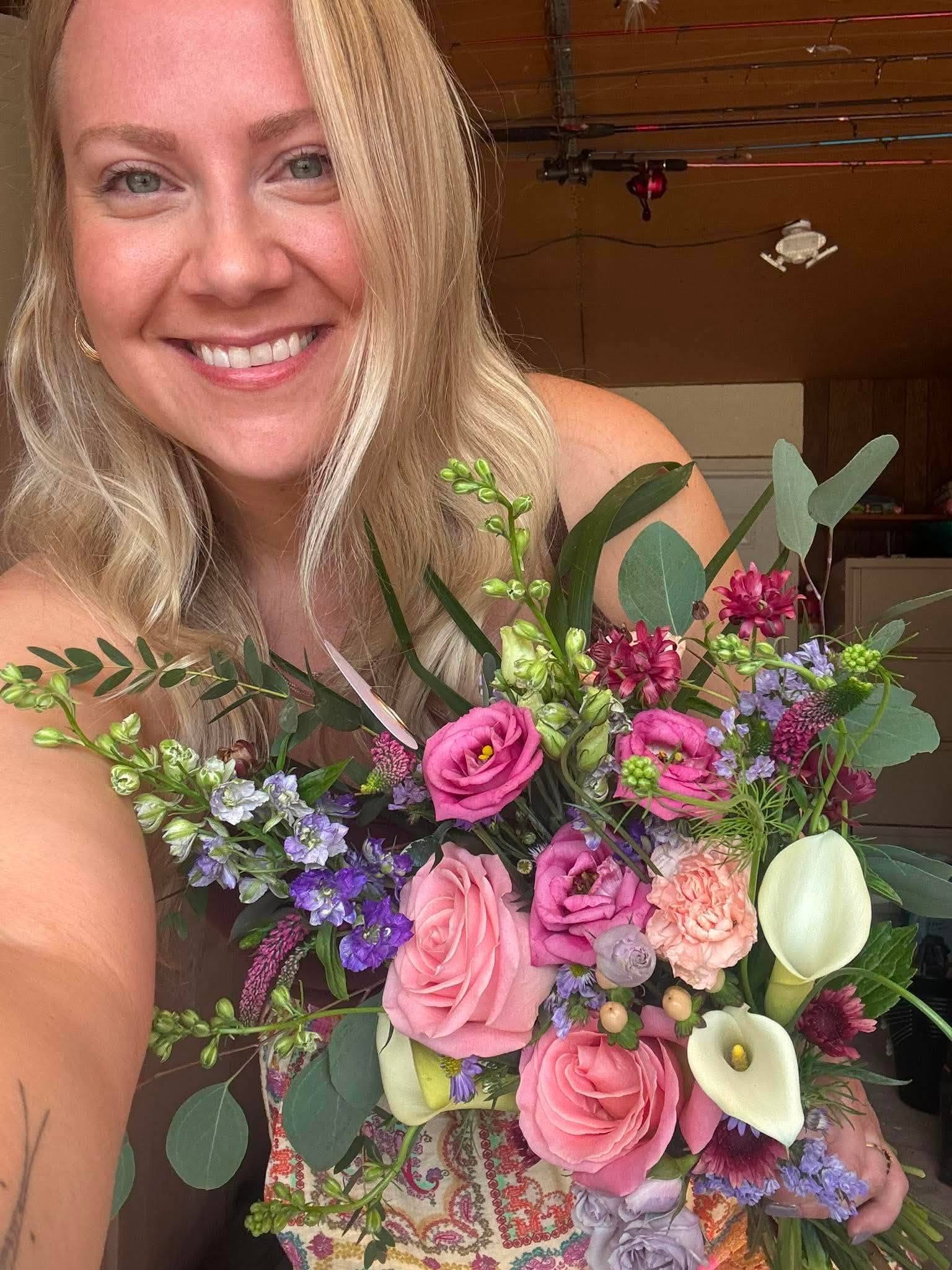 Smiling woman holding a colorful bouquet of flowers, including pink roses, white calla lilies, and purple blooms.