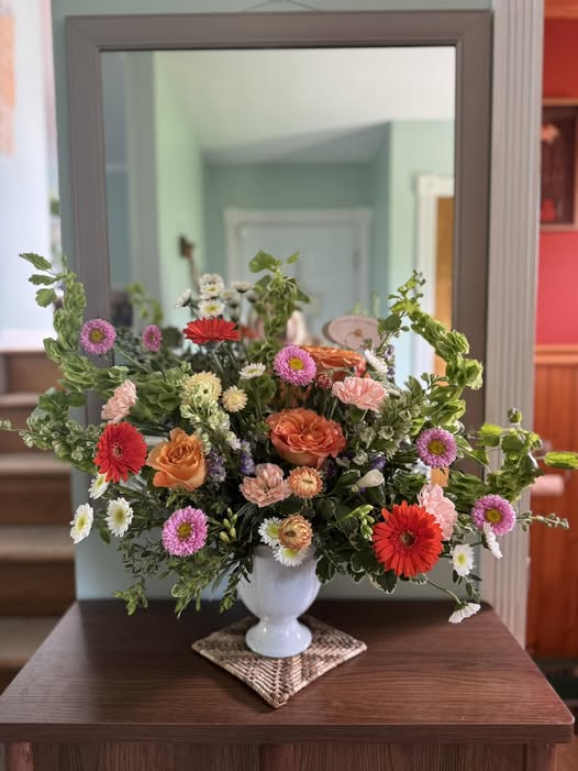 Colorful flower arrangement in a white vase on a wooden table, with a mirror reflecting a room in the background.