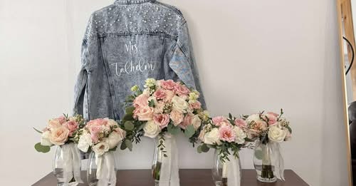 Four glass vases with pink and white roses on a table, denim jacket hanging on the wall behind, with writing on it.