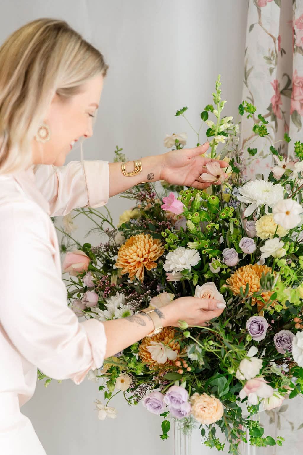 Woman arranging a large colorful flower bouquet indoors, with a floral curtain in the background.