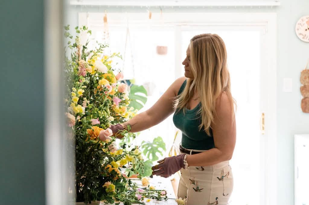 Woman arranging flowers in a bright room with sunlight, wearing a teal top and patterned pants, holding gloves.
