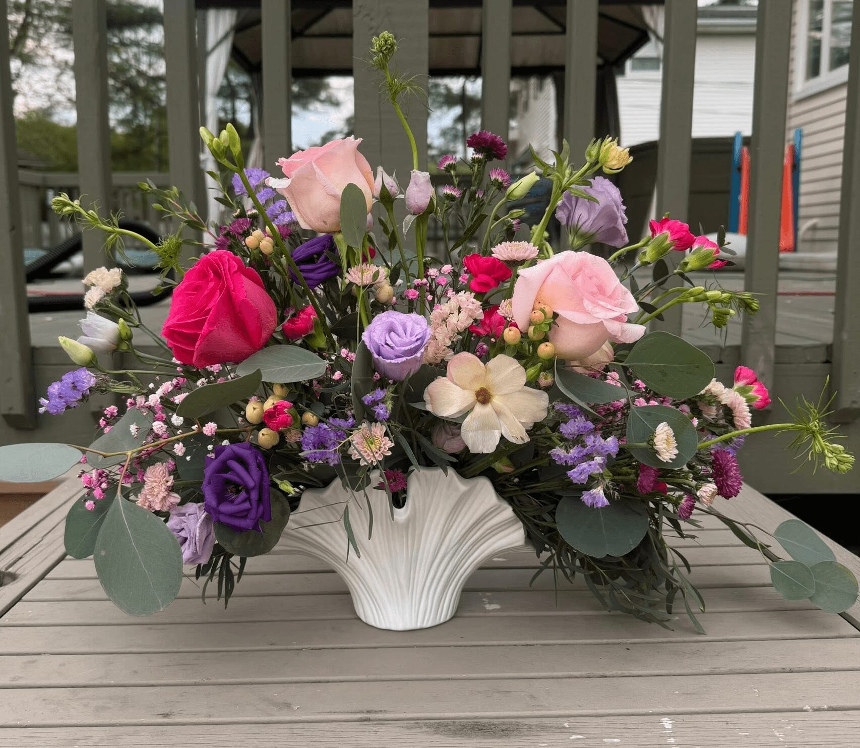 Flower arrangement with pink, purple, and white flowers in a white vase on a wooden table outdoors.