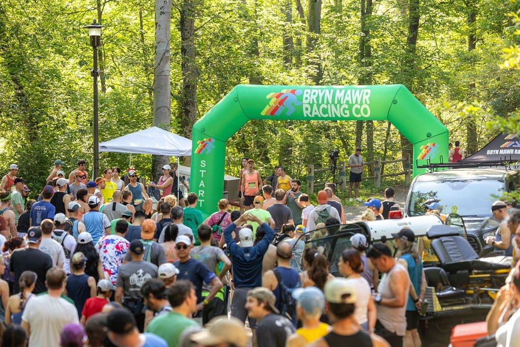 Crowd gathered at start line of outdoor race event in a wooded area with green trees, under a green inflatable arch.