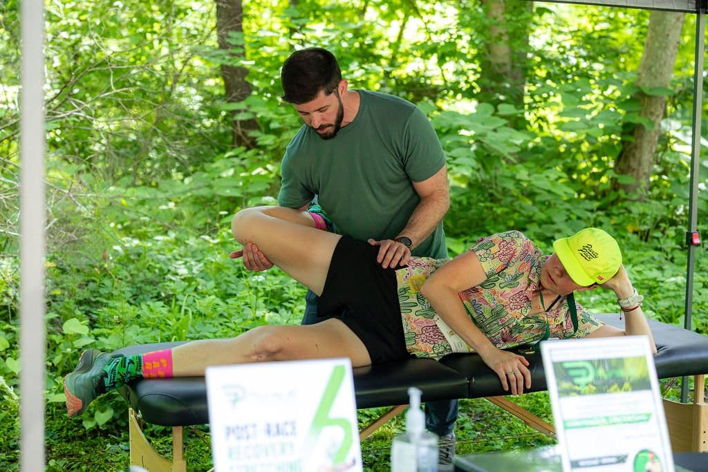 Person lying face down on a massage table outdoors, another person standing beside, in a green forest setting.