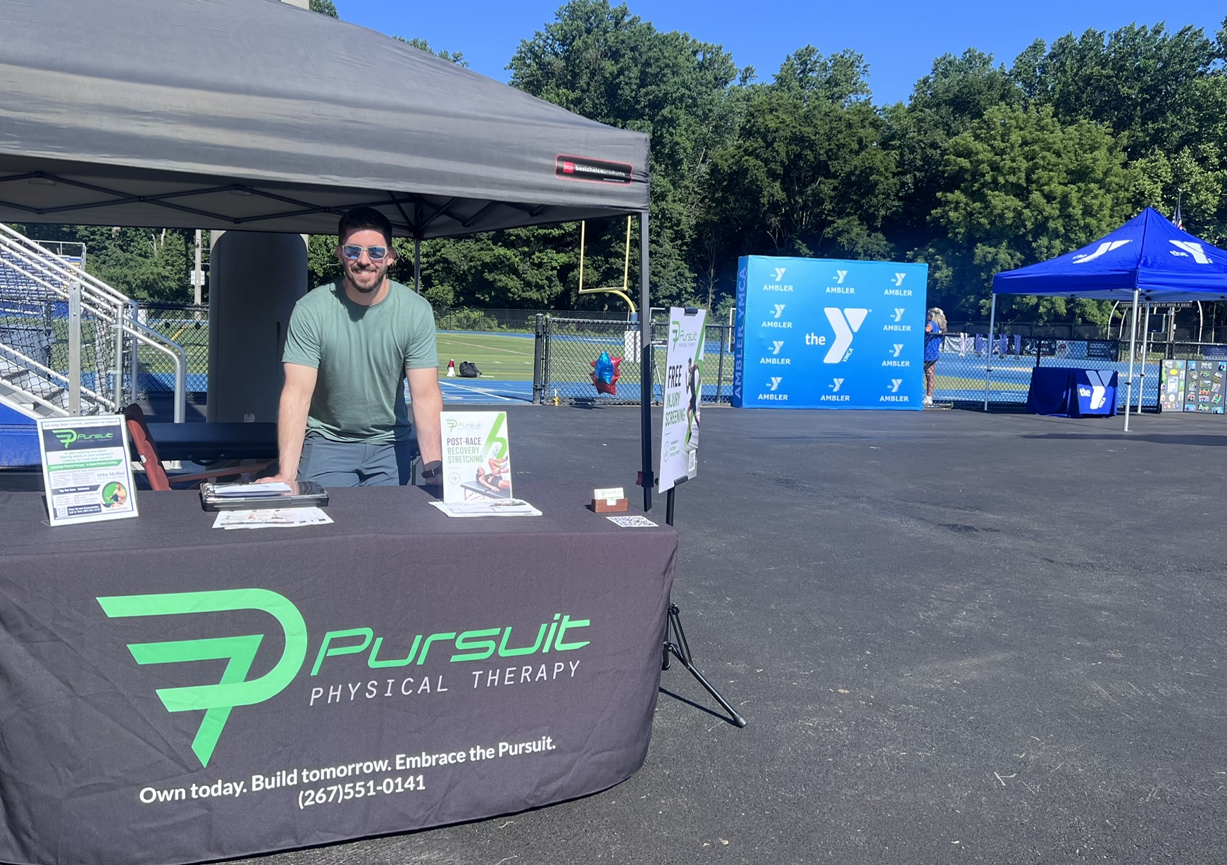 Man standing behind a Pursuit Physical Therapy booth outdoors with trees and tents in background.