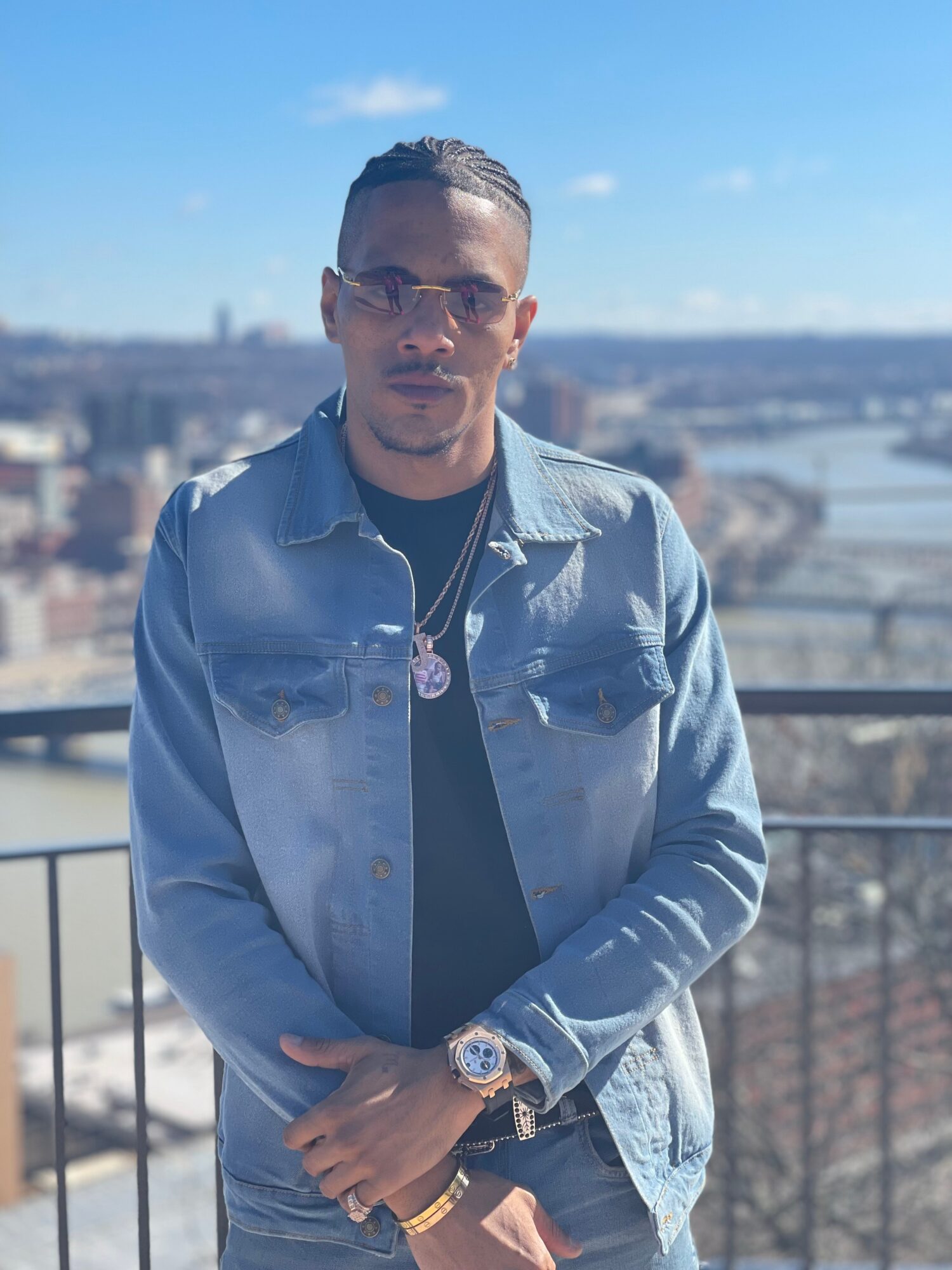 Man wearing sunglasses and denim jacket standing outdoors with cityscape background