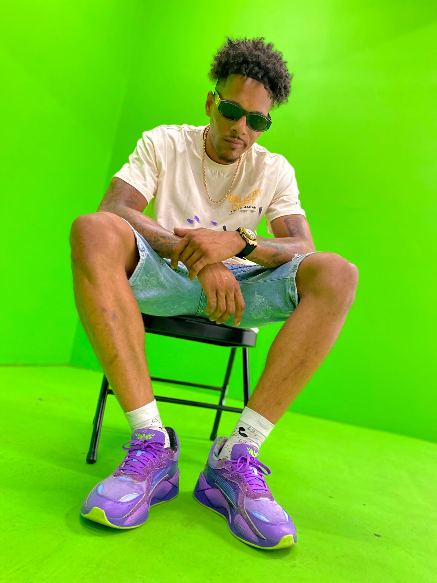 Young man with sunglasses sitting on a chair against a green background, wearing a white shirt, shorts, and purple sneakers.