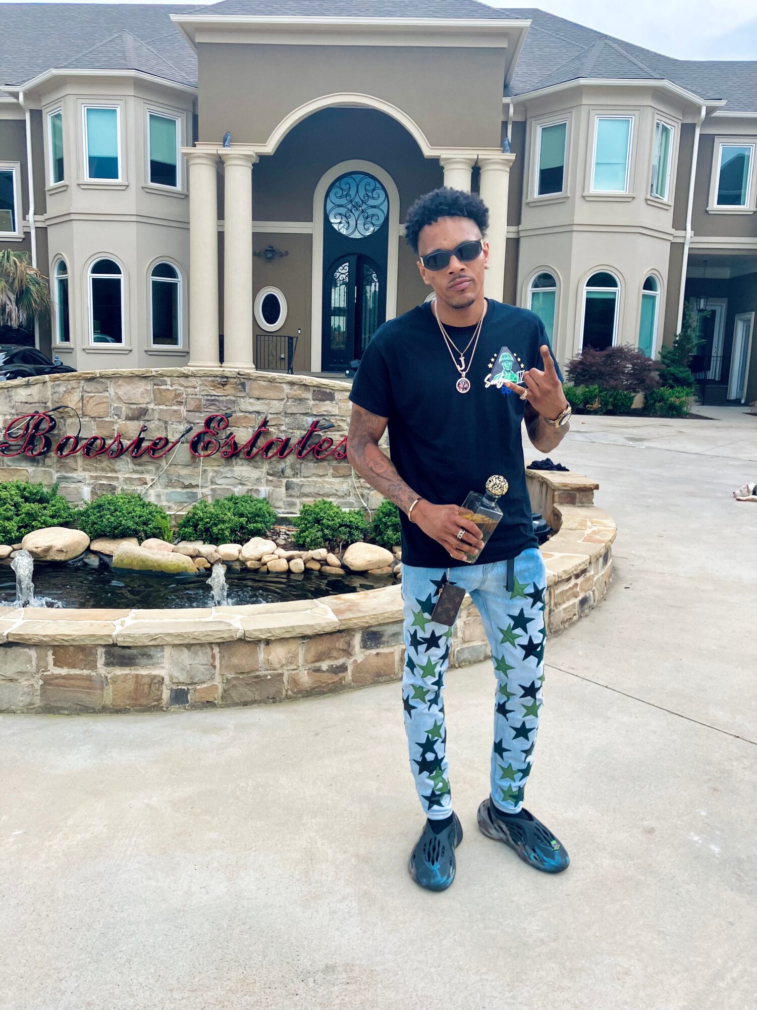 Young man standing in front of a large house with a fountain, wearing sunglasses, black shirt, and star-patterned pants.