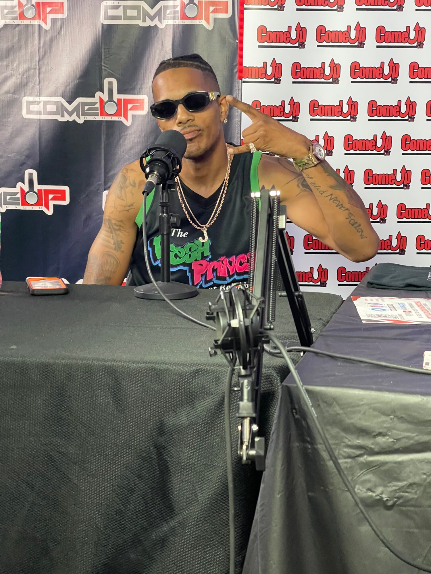 Person wearing sunglasses and a black shirt, speaking into a microphone at an event with a branded backdrop.