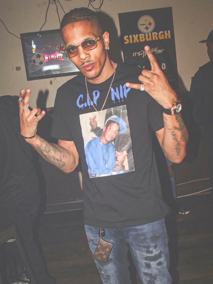 Young man with glasses and tattoos making hand gestures, wearing a black T-shirt with a photo print, in a club setting.