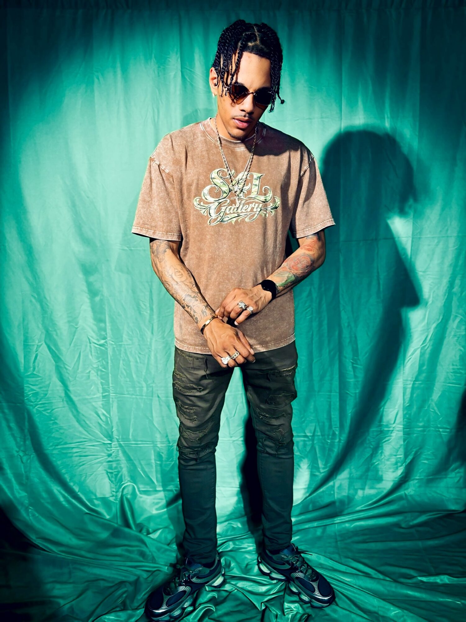 Young man with sunglasses and tattoos stands against a teal backdrop, wearing a brown T-shirt and black pants.