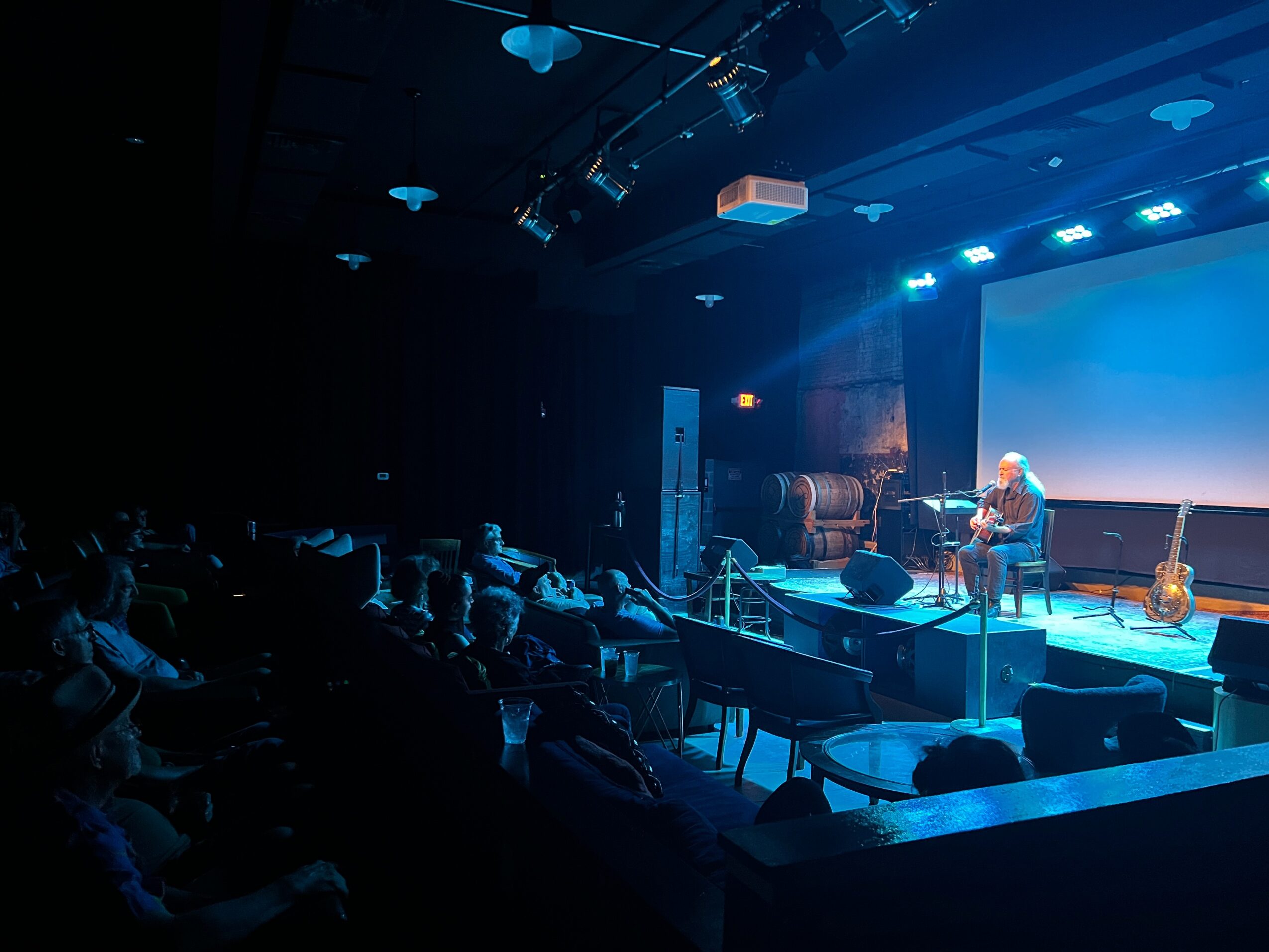 Stage with two people, musical instruments, and a large screen, audience seated in a darkened room.