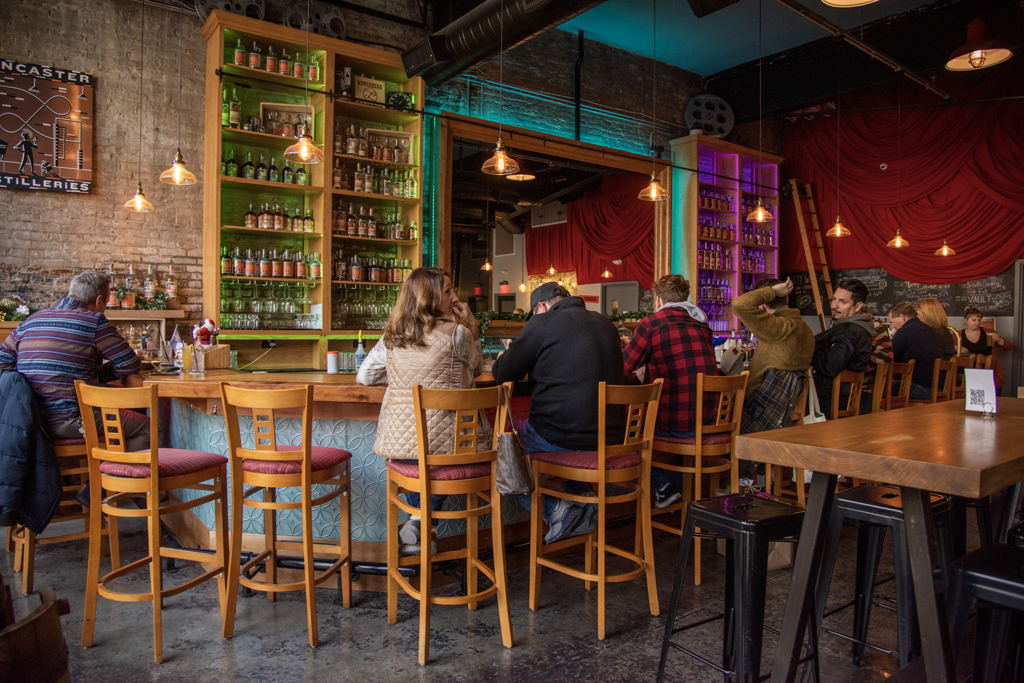 People sitting at a bar counter in a cozy, colorful pub with shelves of bottles and warm lighting.