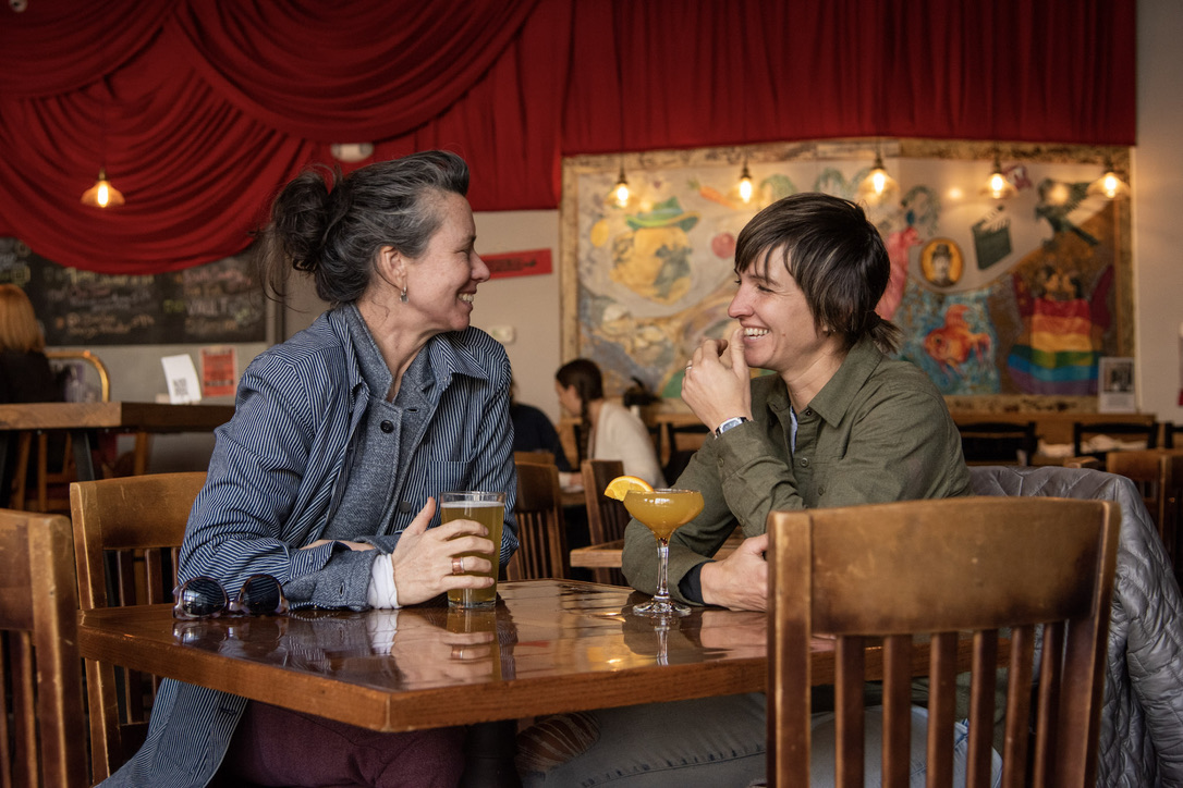 Two women sit at a table in a lively bar, smiling and talking, with drinks and a yellow cocktail on the table.