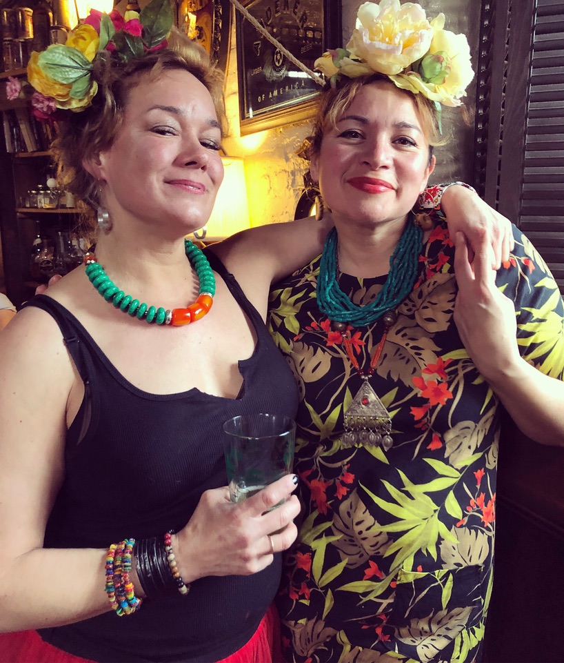 Two women with floral headpieces and colorful clothing, smiling and posing together indoors, one holding a glass.