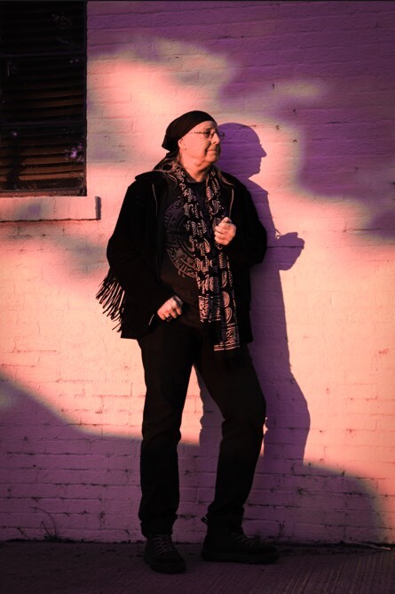 Woman standing against a wall with colorful lighting, wearing glasses, a scarf, and a coat, holding a bag strap.