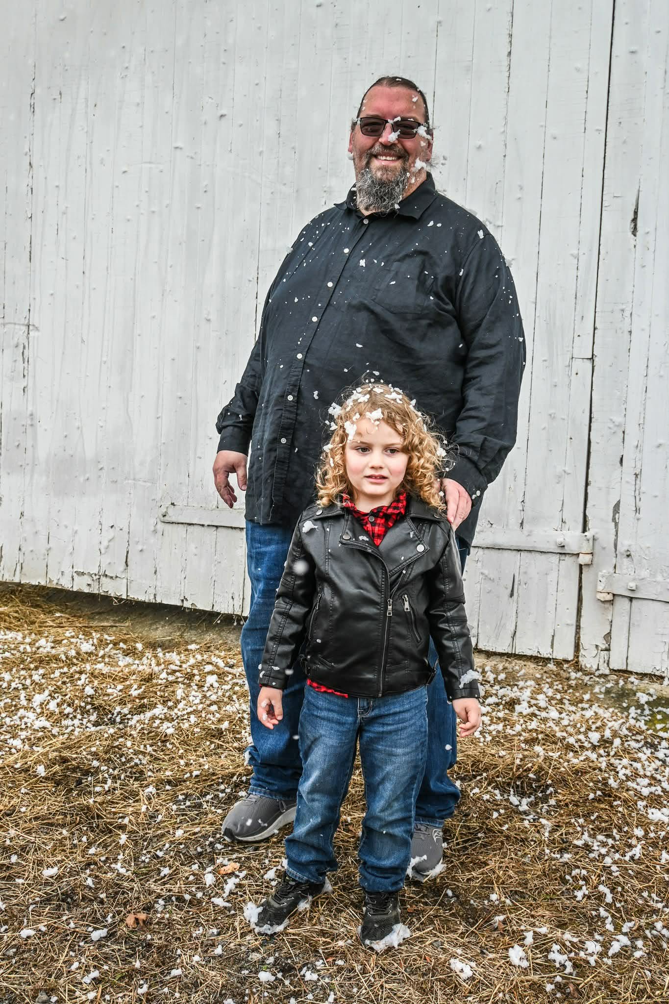 A man and a young girl stand outdoors in front of a white wooden wall, smiling, with snow on the ground.
