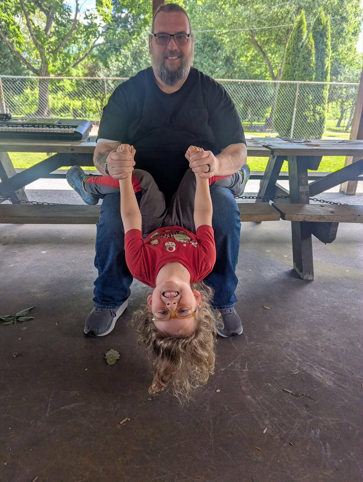 Adult man sitting on a bench holding a smiling child's hands upside down, child hanging upside down, outdoors with trees and picnic tables