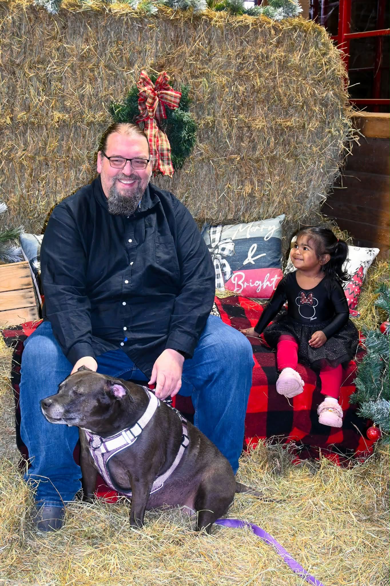 Man with glasses and beard sitting on hay bale, young girl sitting nearby, dog in front, Christmas decorations behind.