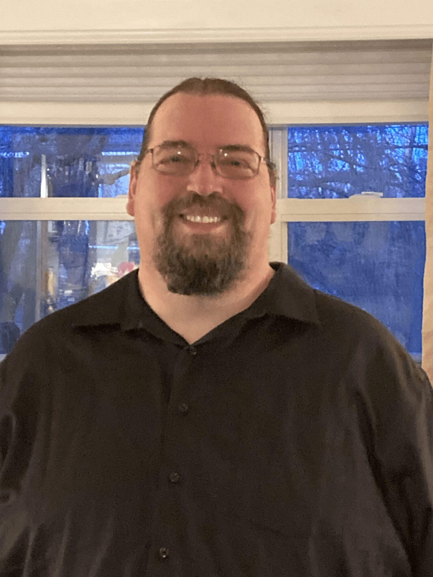 Smiling man with glasses and beard wearing a black shirt indoors, with a window showing trees outside.