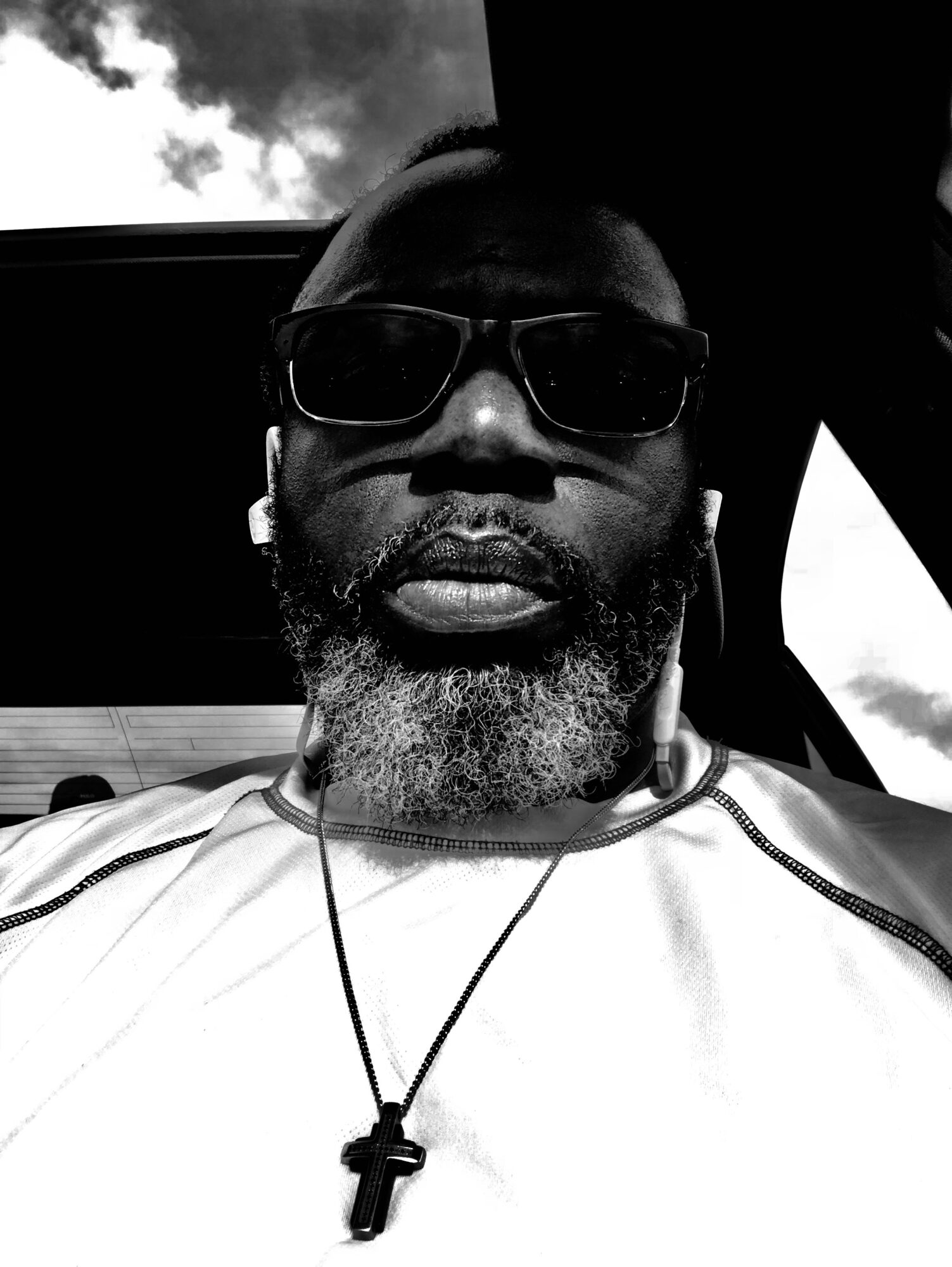 Man with sunglasses and a beard, wearing a necklace with a cross, sitting inside a vehicle, with a cloudy sky visible outside.