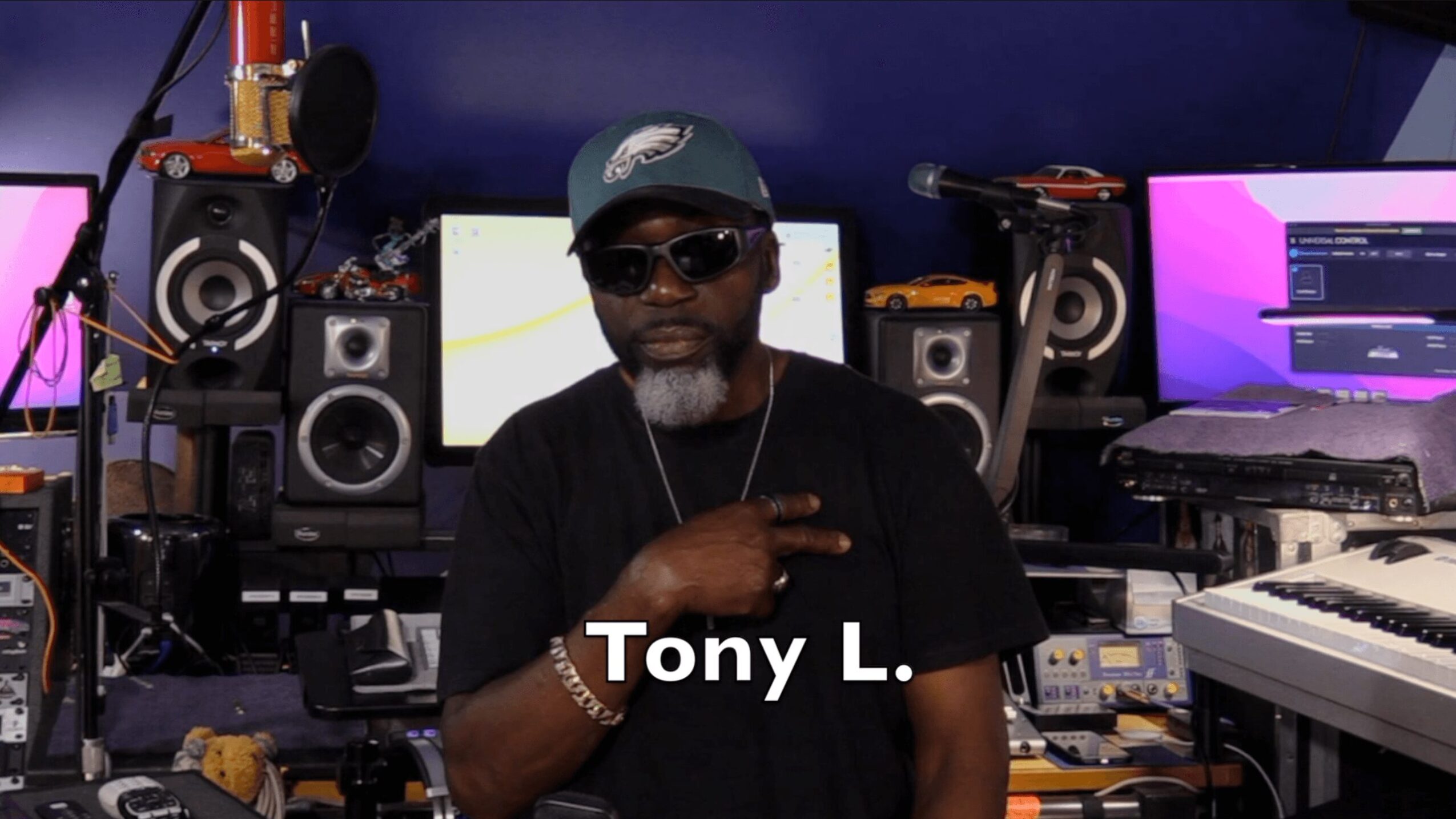 Man wearing sunglasses and a cap in a music studio with equipment and screens behind him.