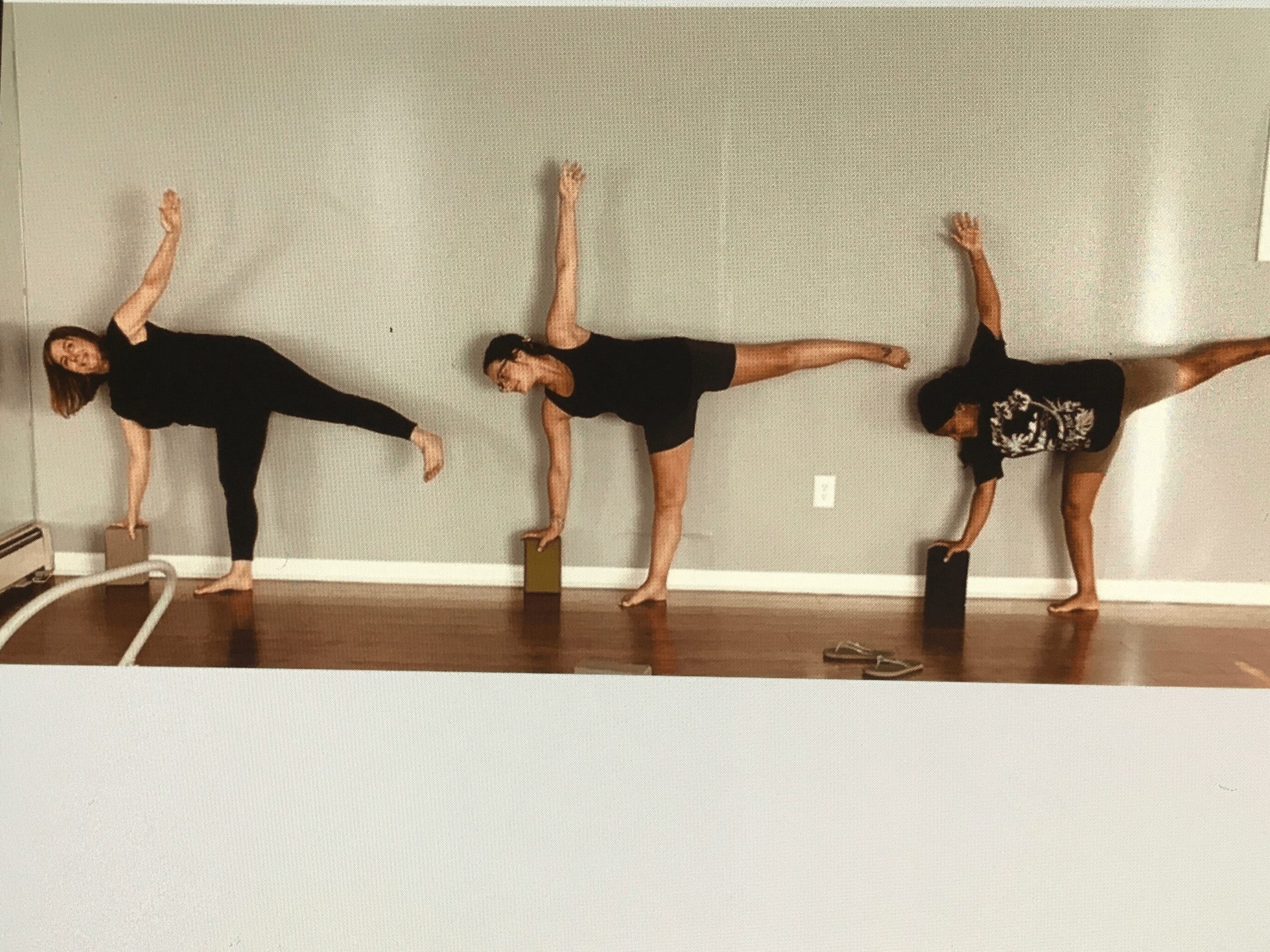 Three people practicing yoga poses against a gray wall, each balancing on one leg with arms raised or extended.