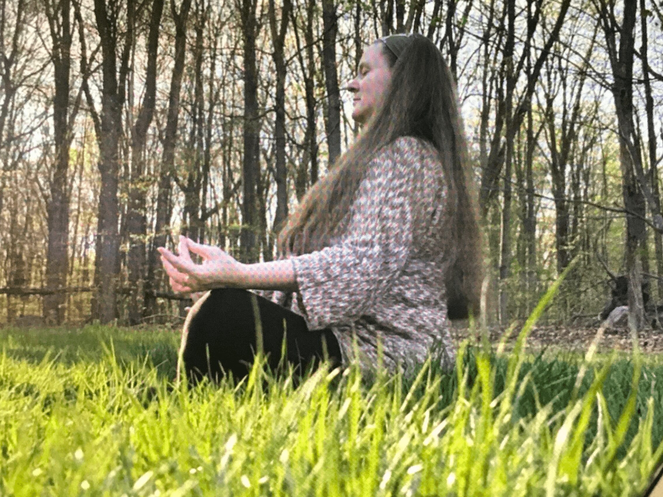 Woman sitting cross-legged outdoors in a forest, meditating with eyes closed, surrounded by trees and grass.