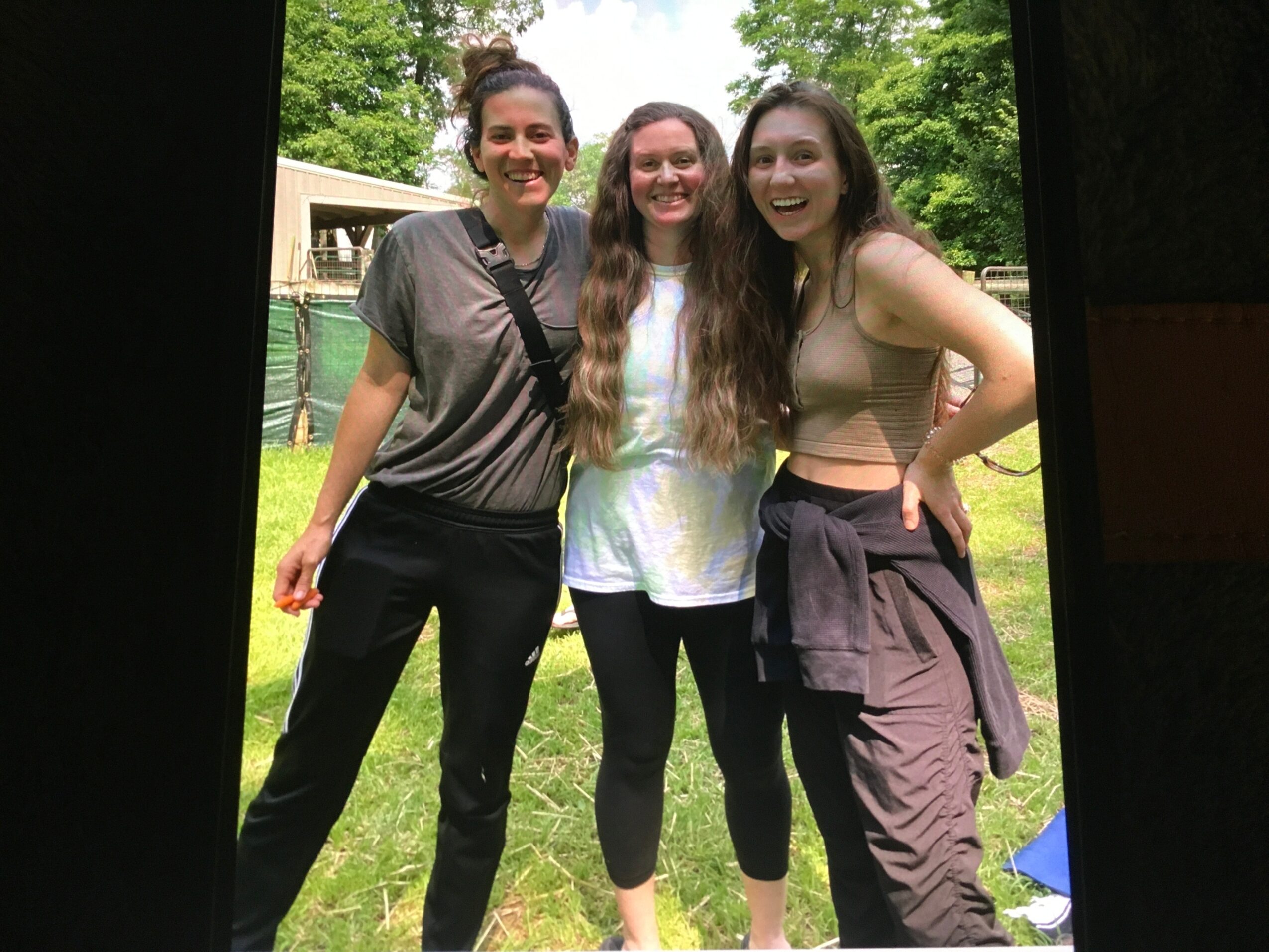 Three women standing outdoors on grass, smiling, with trees and a building in the background.