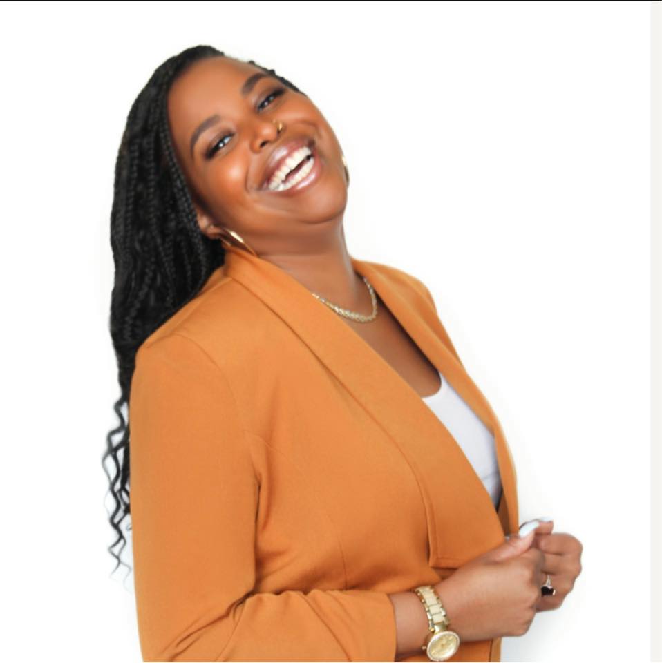 Smiling woman with long curly hair wearing an orange blazer and white top, posing with hands clasped, against a white background.