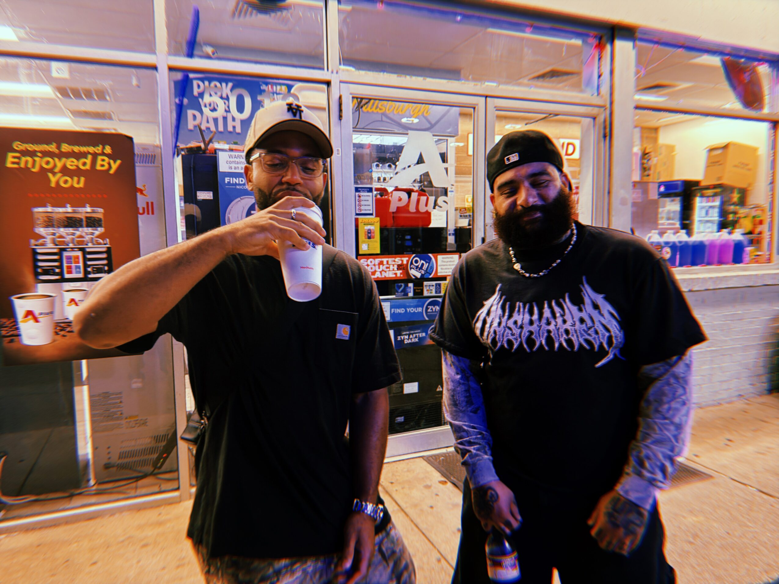Two men standing outside a store, one holding a drink, the other with a bottle, both smiling. Store window behind them.