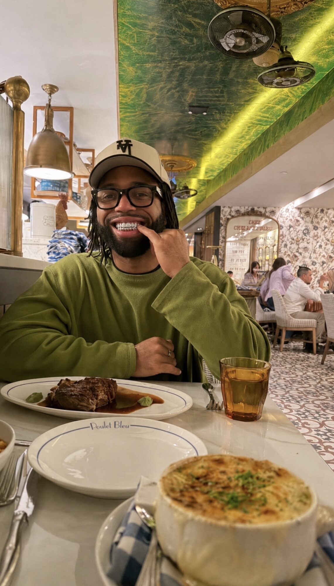 Smiling man wearing glasses and a cap, sitting at a restaurant table with food and drink, background with decorative ceiling and other diners.