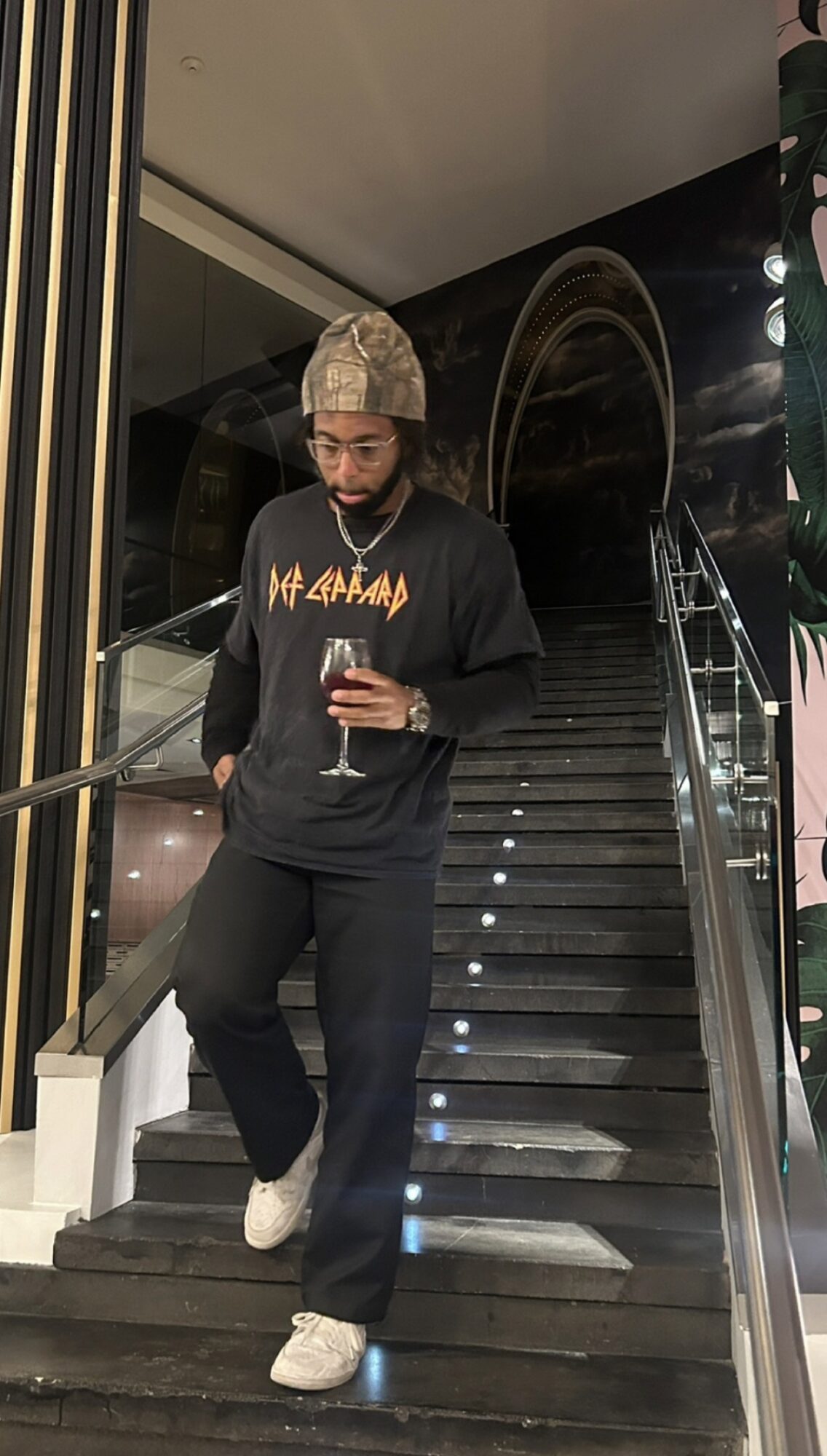 Person in black outfit and camouflage hat walking down stairs holding a drink, indoors at night.